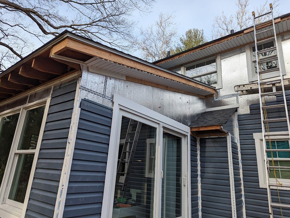 Home exterior under construction; silver insulation, blue siding, white window trim, ladder.