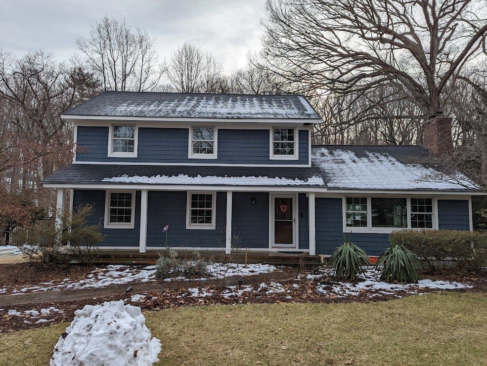 Two-story blue house with white trim, porch, and snow-covered roof, set in a winter landscape.