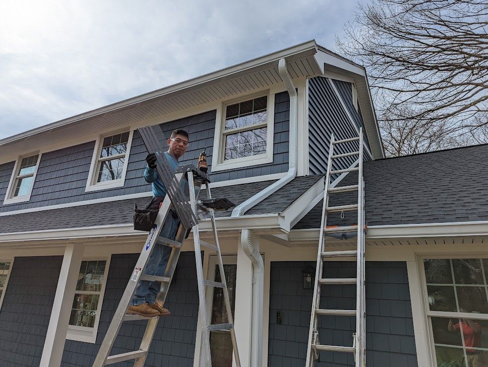 Man on ladder installing siding on a two-story house with blue siding and white trim on a sunny day.