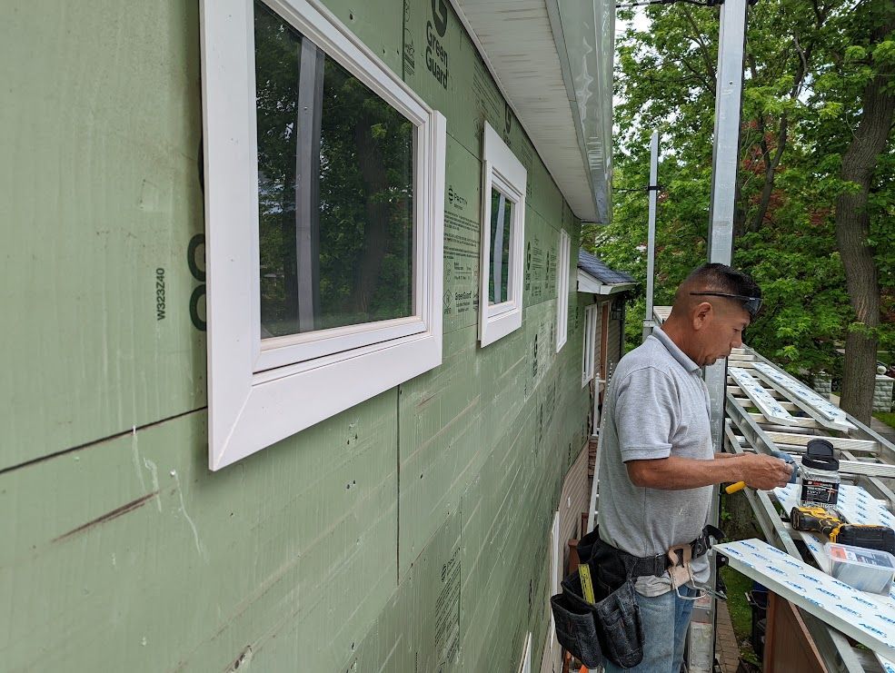 Man installing siding on a house, standing on a scaffold. Windows and green insulation are visible.