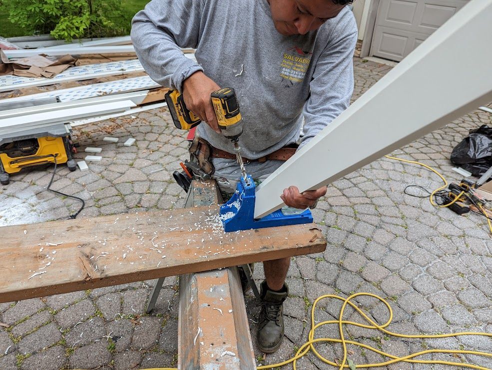Person using a drill to secure white trim with a blue jig to a wooden board outdoors.