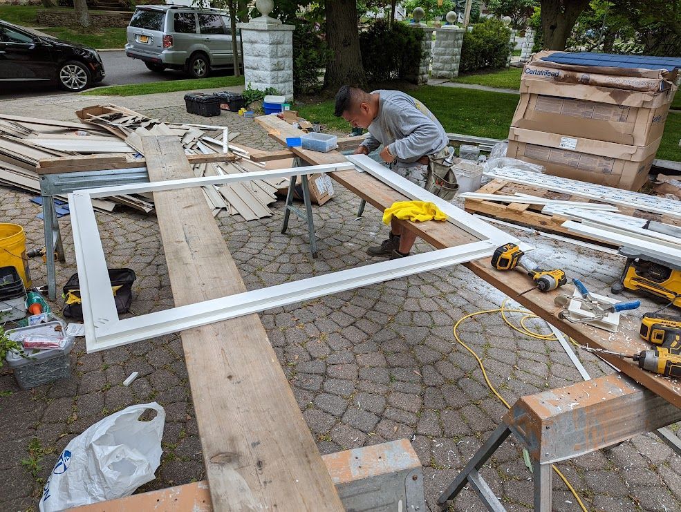 Man working on a white window frame outdoors on a wood plank, tools and materials scattered around.