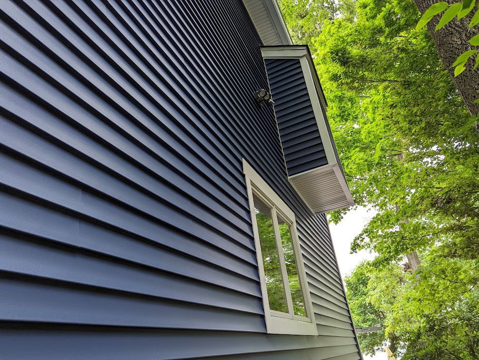Blue clapboard siding on a house with white trim window, with green foliage in the background.