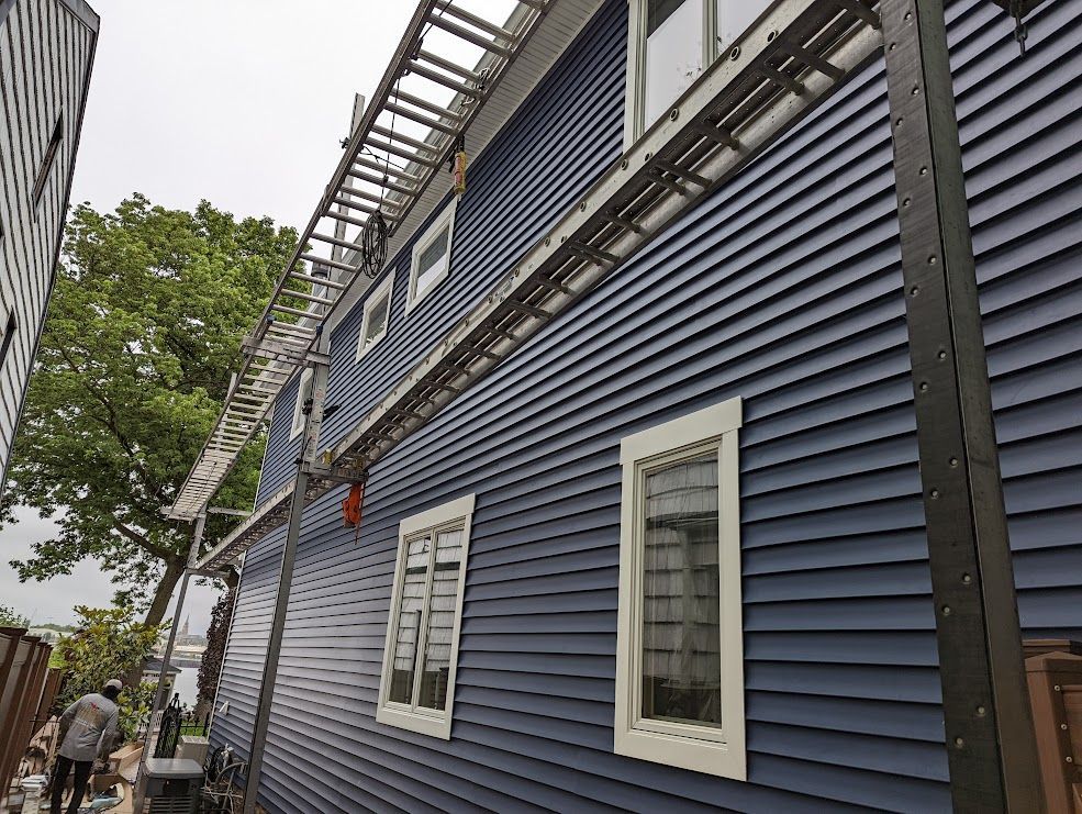 House with blue siding, two ladders leaned against the wall, window trim, and a cloudy day.