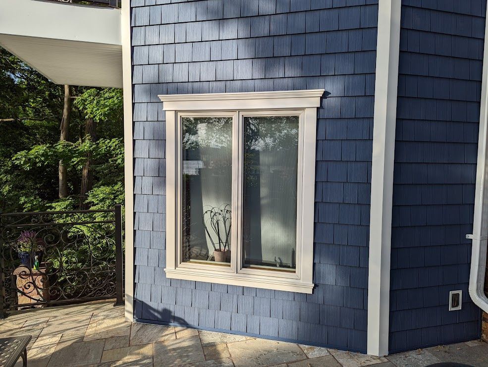 Blue shingle siding with a window framed in cream. Exterior view with lush greenery and a stone patio.