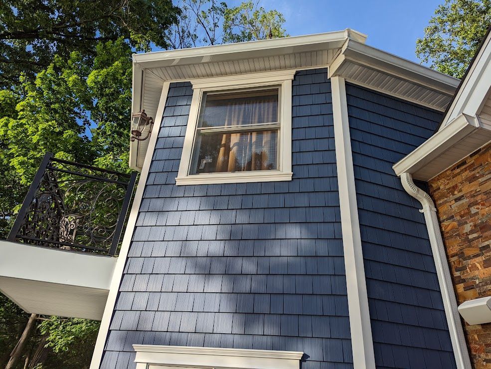 Blue-shingled house with a window and white trim. Balcony with ornate railing, trees in the background.