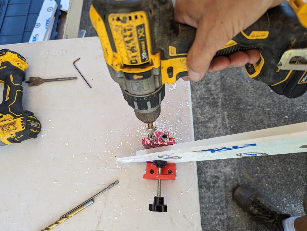 A person using a yellow drill to bore a hole into a white board clamped to a red vise.