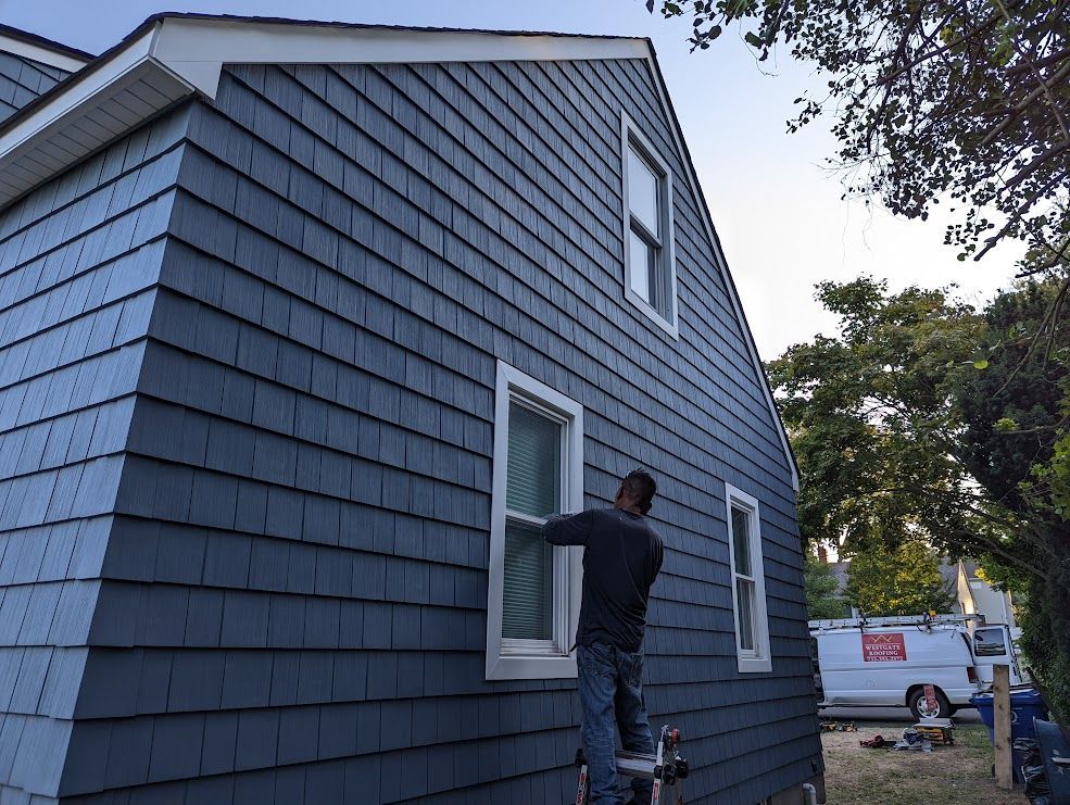 Man on a ladder working on dark blue shingle siding of a house with white window frames.