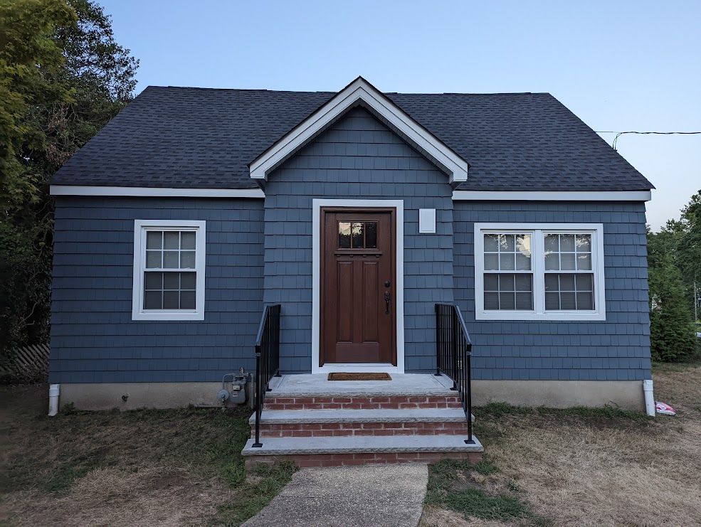 Blue-sided cottage with brown door, white-framed windows, brick steps, and a dark roof.