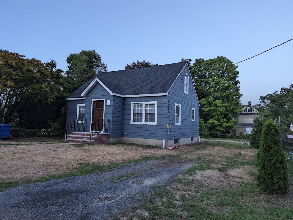 Blue house with dark roof and white trim, sitting on dry, grassy land. Gravel driveway leads to the house.