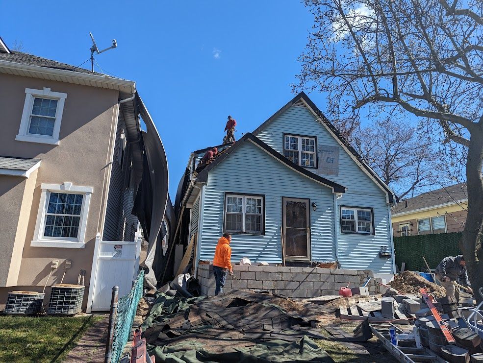 Two-story houses, one blue being worked on; person in orange near foundation, others on roof, blue sky.