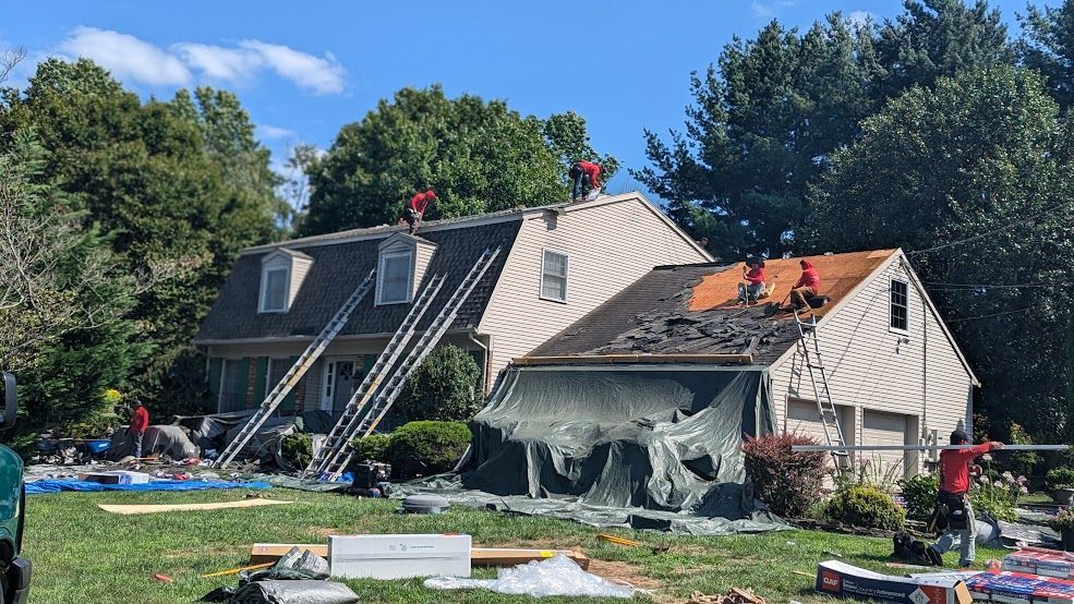 Roofers removing shingles from a house roof on a sunny day. Ladders, tools, and materials are visible.
