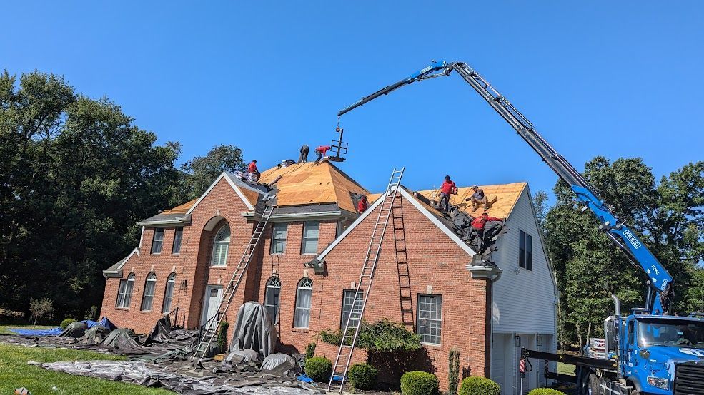 Roofing crew working on a two-story brick house, crane lifting materials. Blue sky, sunny day.
