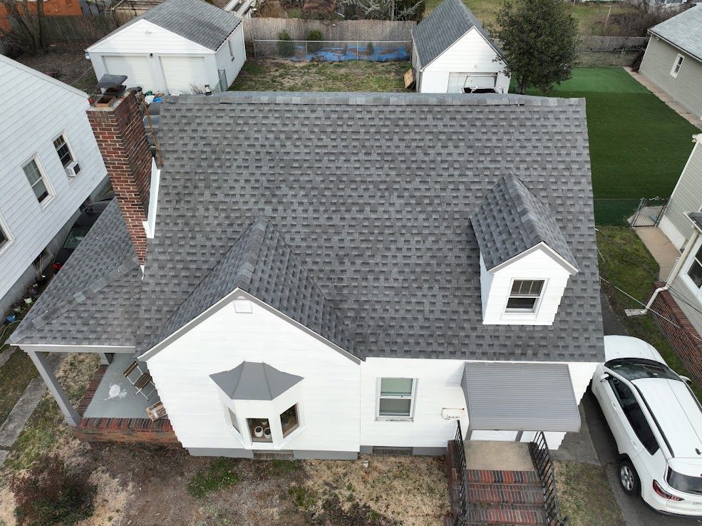 Aerial view of a white house with a gray shingled roof, chimney, and dormer windows, with a white SUV parked in the driveway.
