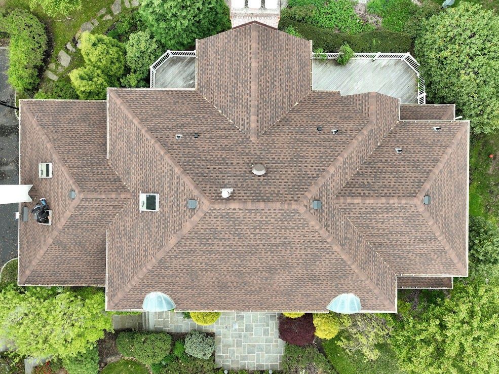 Overhead view of a house with a brown shingled roof, surrounded by green trees and landscaping.