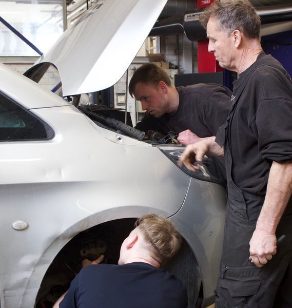 Tre mekanikere arbejder på en sølvfarvet bil med motorhjelmen oppe i en garage.