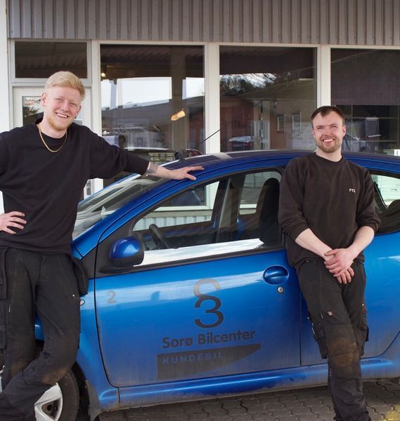 To mænd står ved siden af ​​en blå bil. Den ene læner sig smilende op ad den, den anden smiler, foran en bygning.