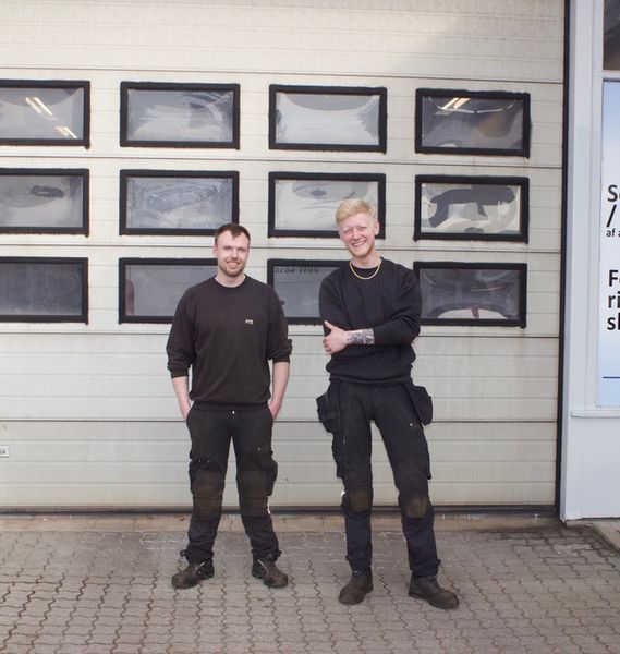 To mænd står foran en garageport. Begge iført arbejdstøj og smiler.