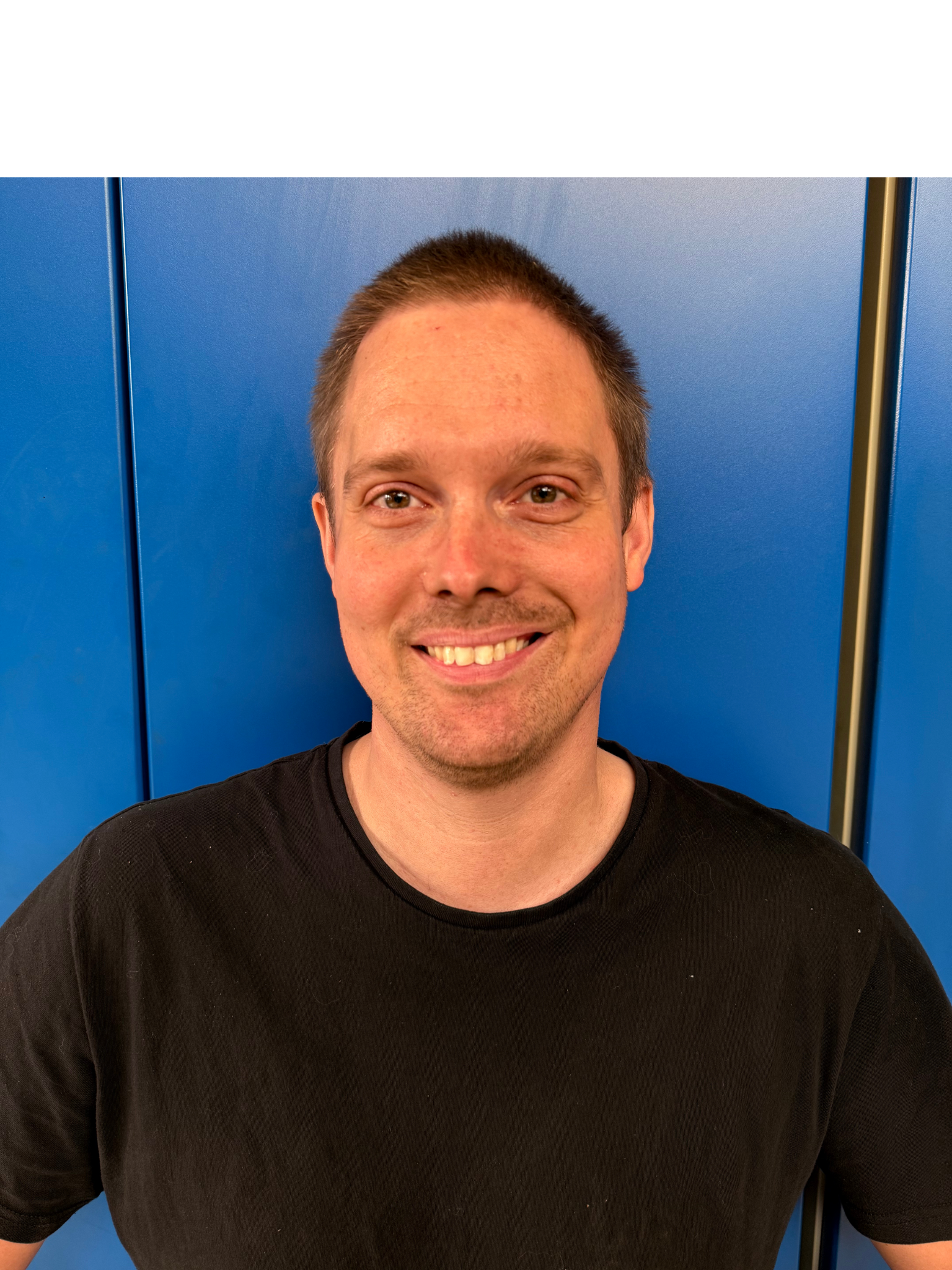 En mand smiler, står foran en blå væg iført en sort skjorte.