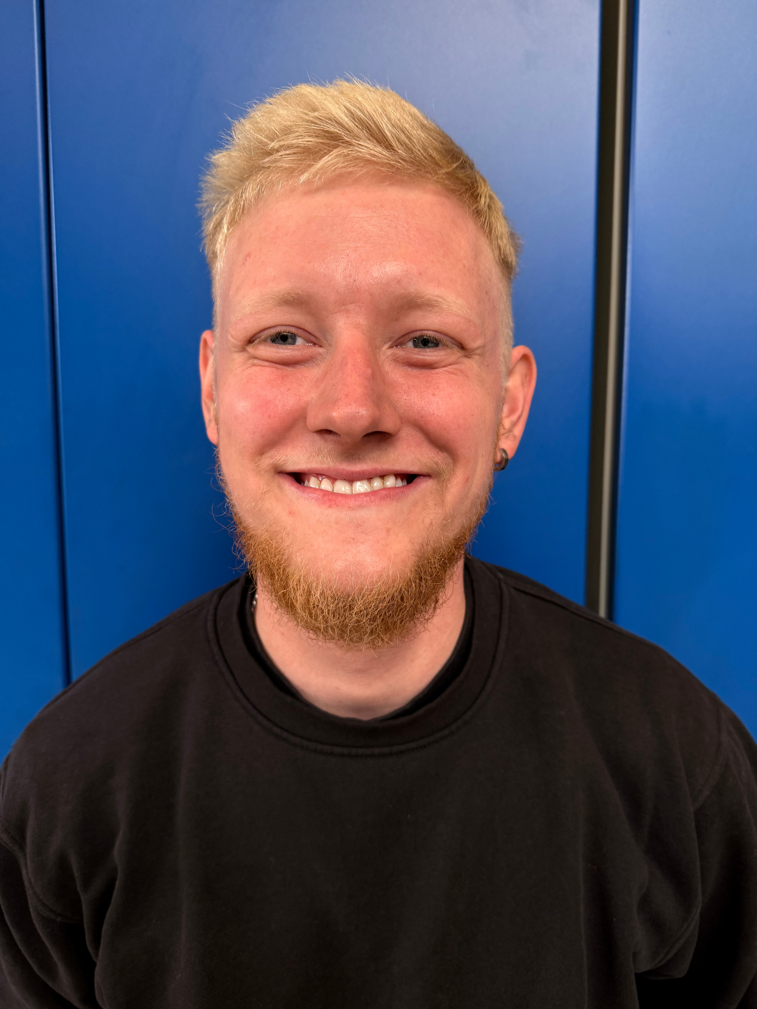 En blond mand med fipskæg smiler til kameraet. Han er iført en blå skjorte og en guldhalskæde foran blå paneler.