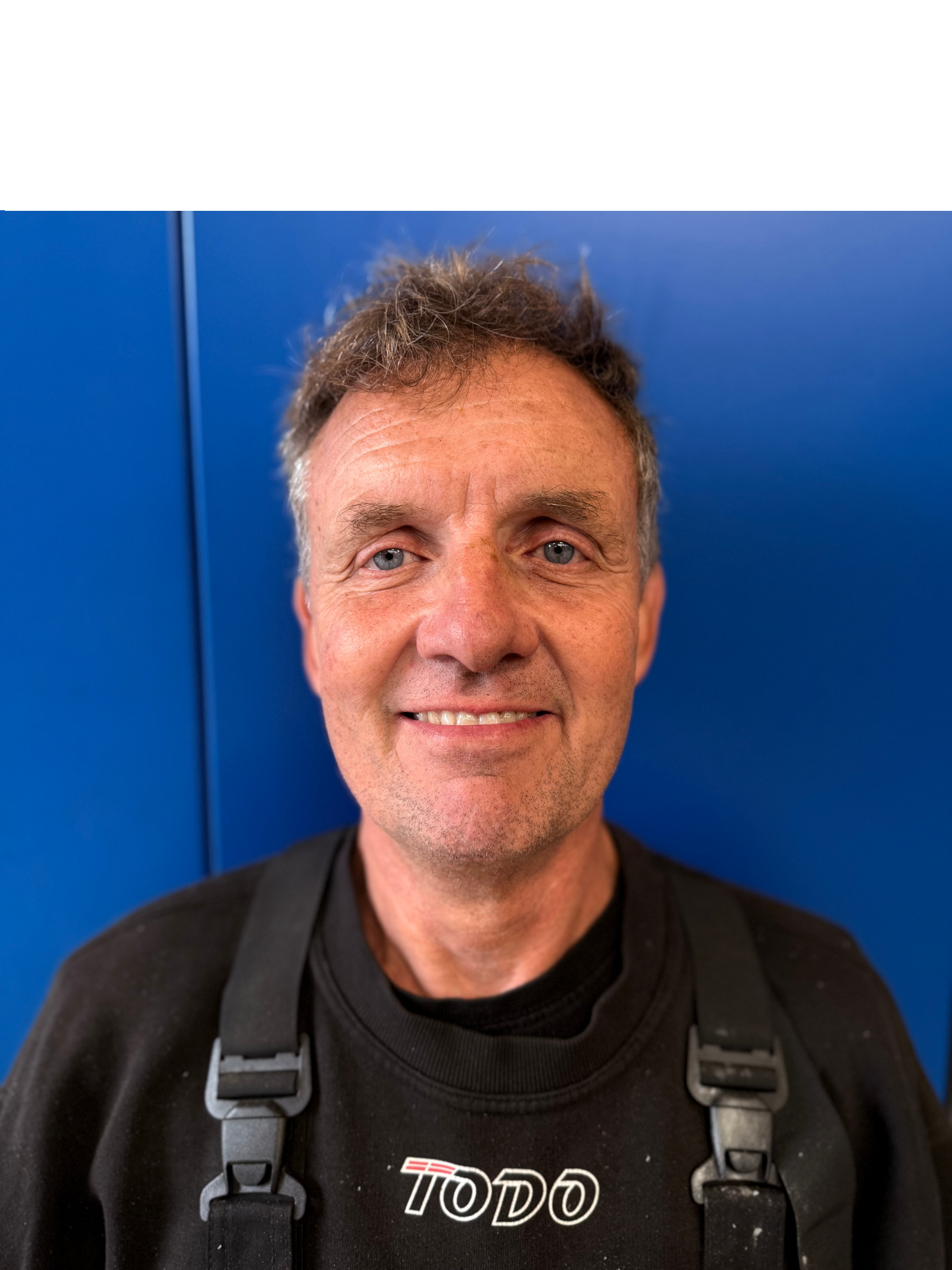 Mand med lys hud og kort brunt hår smiler, stående foran en blå væg.