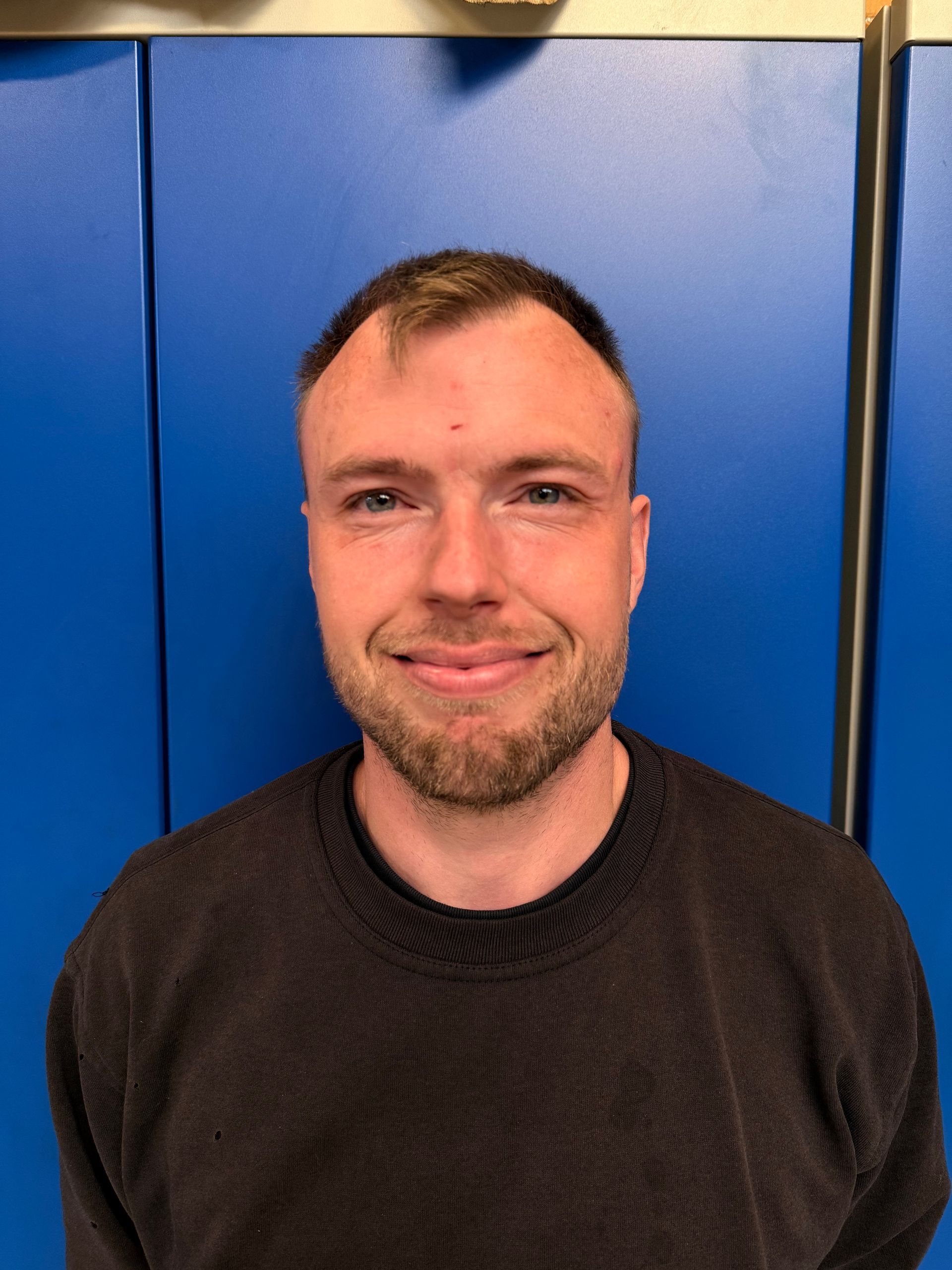 En mand med kort hår og skægstubbe smiler foran en blå væg.