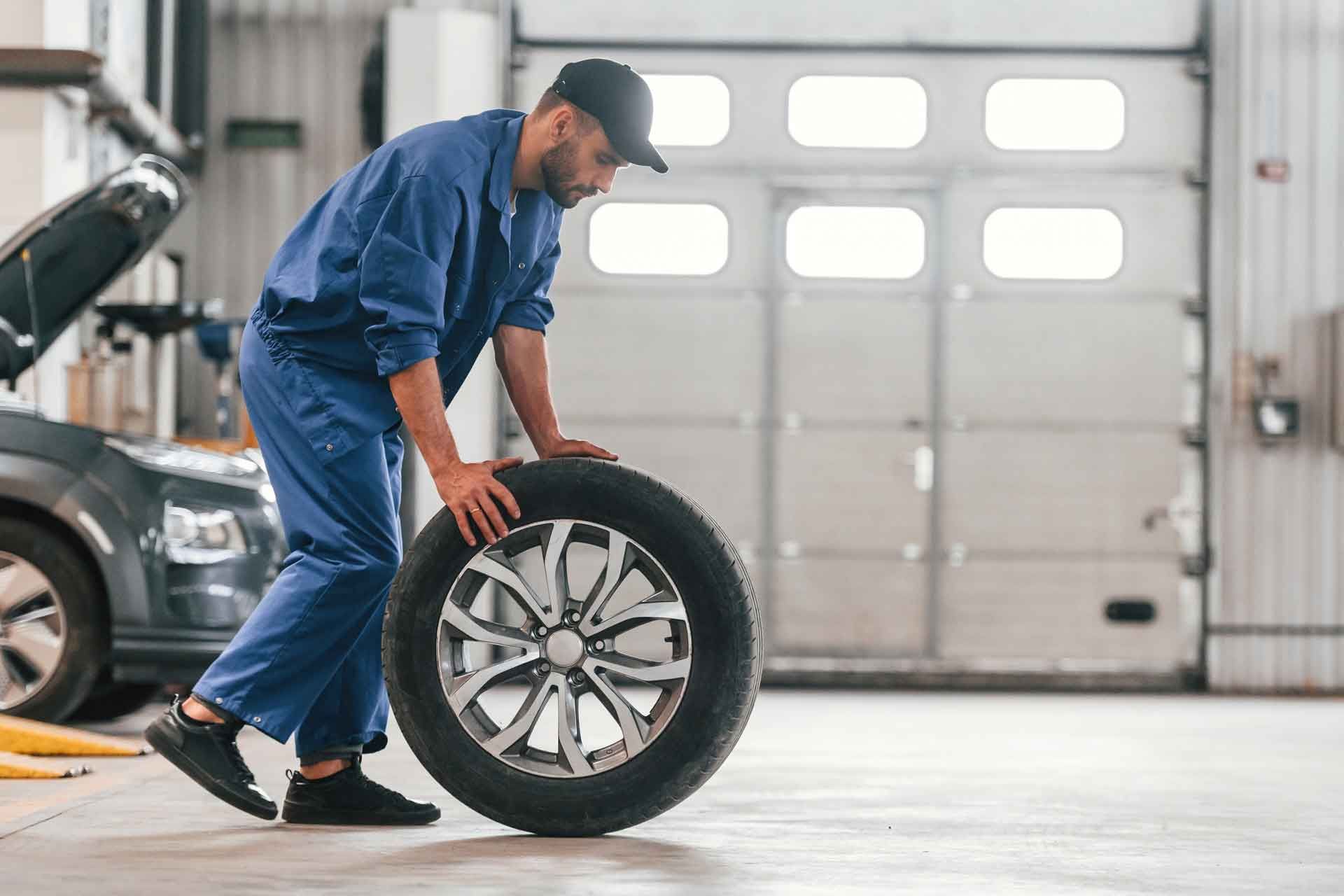 Mekaniker i blå dragt ruller et dæk på garagegulvet med en bil i baggrunden.