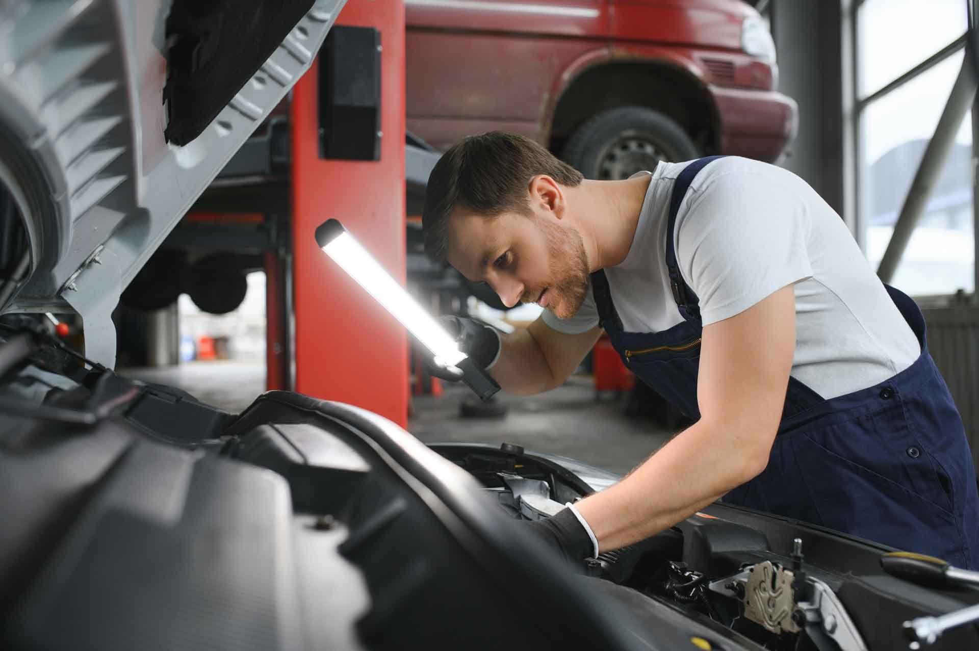 Mekaniker arbejder på en bilmotor og bruger en arbejdslampe. Garageindstilling, mand i overalls.