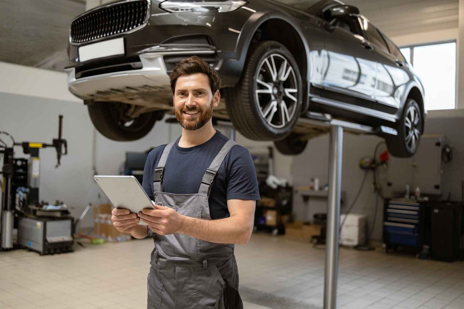 Mekaniker, der holder en tablet, smiler i et autoværksted under en løftet bil.
