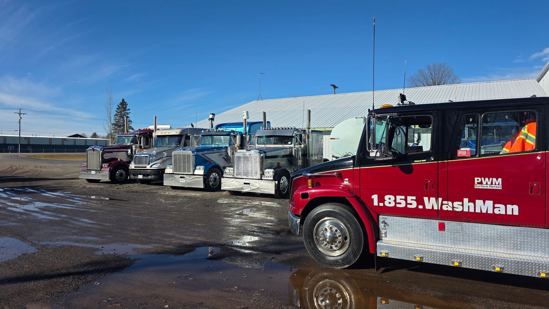 A red WashMan truck parked in a gravel lot next to several parked semi-trucks on a sunny day.