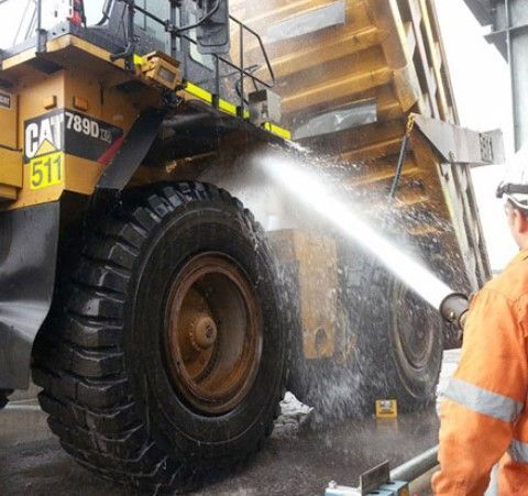 A worker in orange high-visibility gear power-washes a large, yellow Caterpillar 789D mining truck wheel.