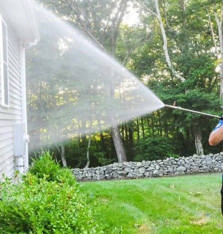 A person uses a pressure washer to clean the light-colored vinyl siding of a house exterior near a green bush.