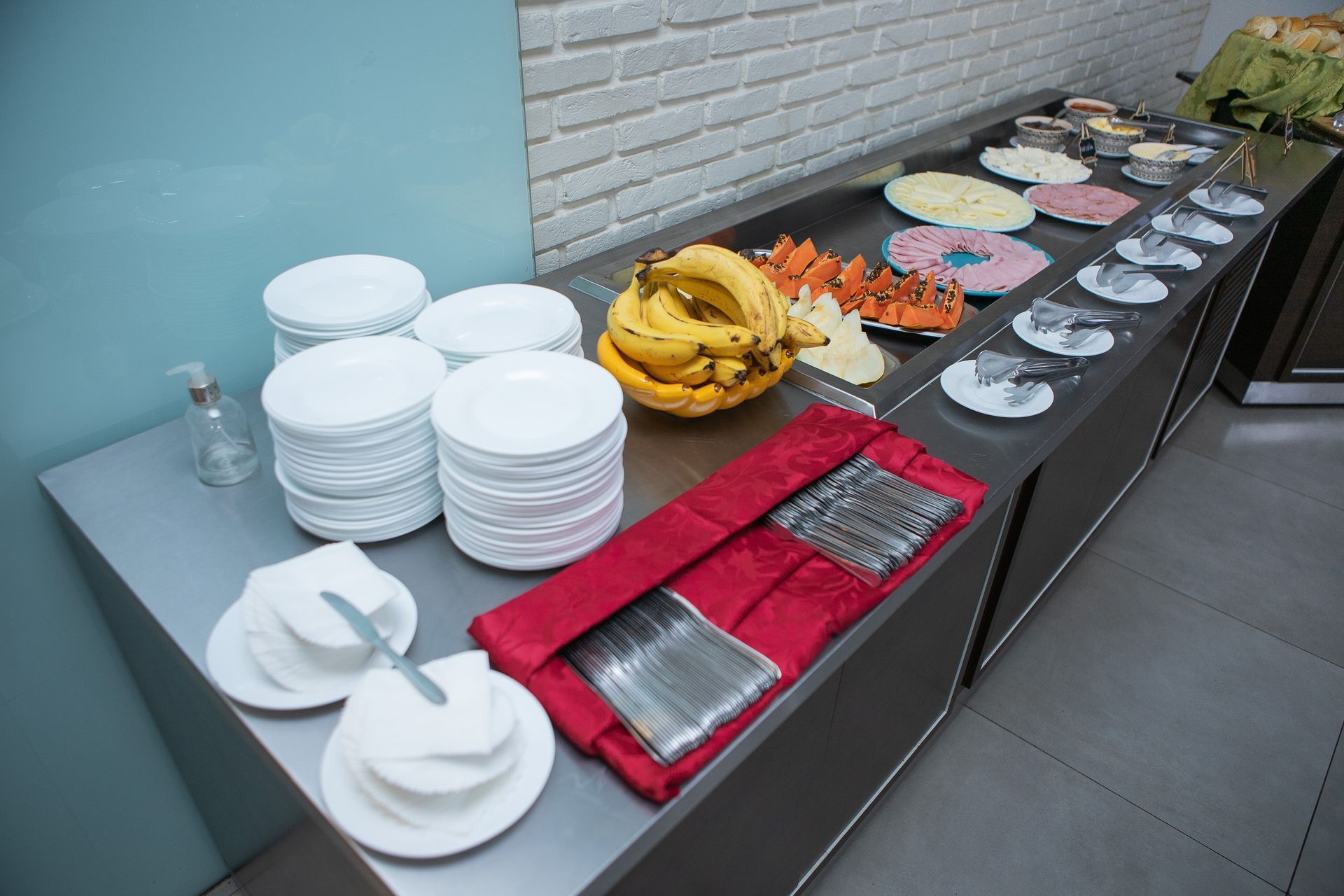 Buffet com diversos itens alimentícios, pratos brancos, talheres e bananas em um balcão de aço inoxidável.