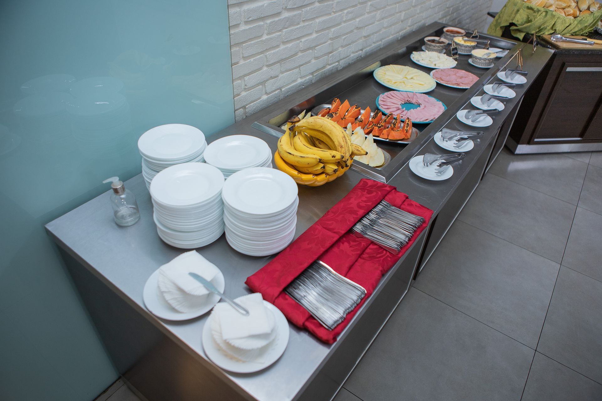 Buffet com diversos itens alimentícios: frutas, pratos e condimentos em um balcão de aço inoxidável.