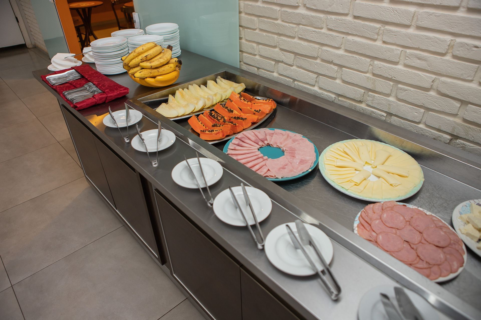 Buffet de café da manhã com frutas, frios e queijos em um balcão de aço inoxidável.