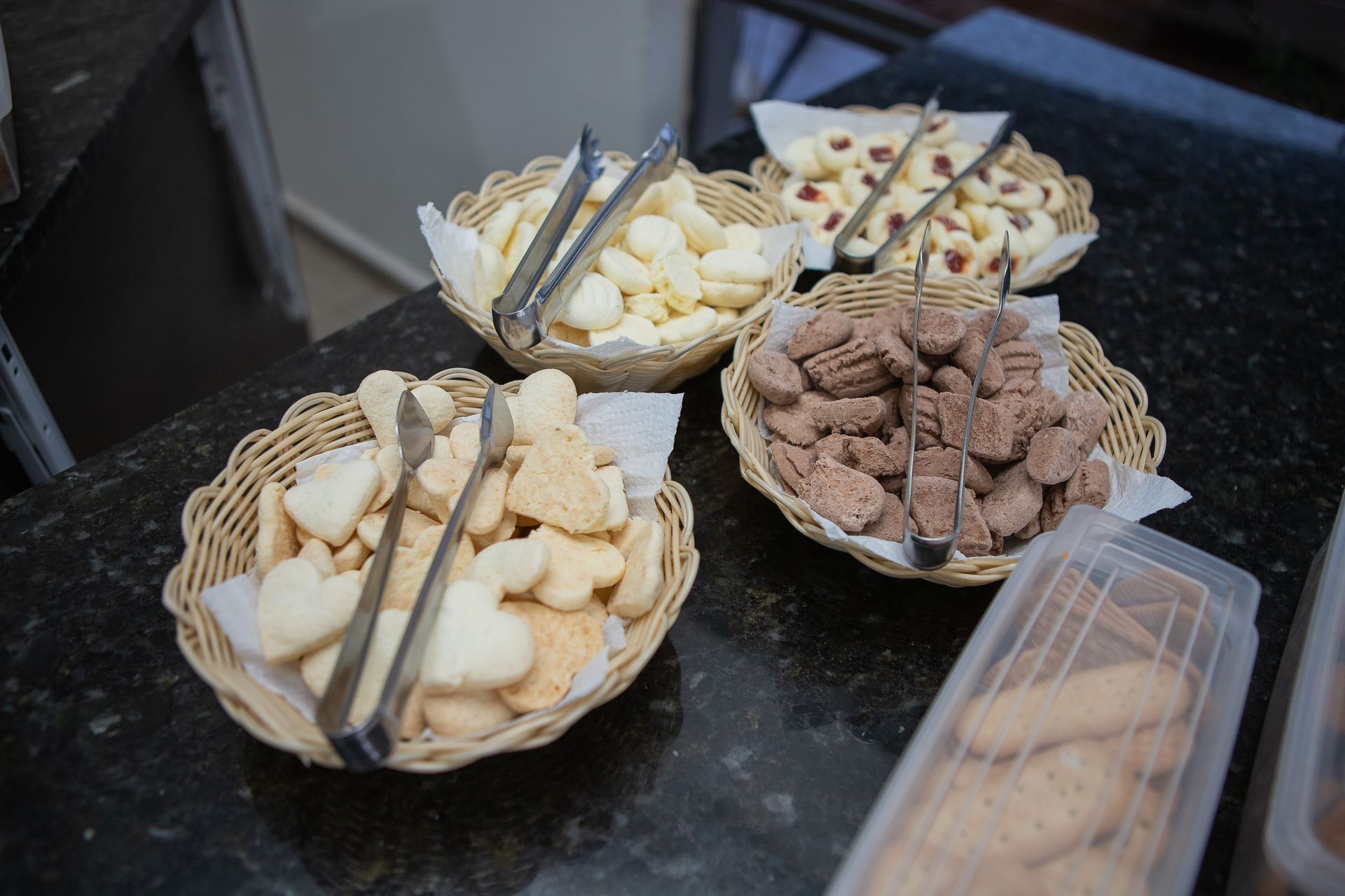 Quatro cestos de vime cheios de biscoitos variados e uma pinça sobre uma bancada escura.
