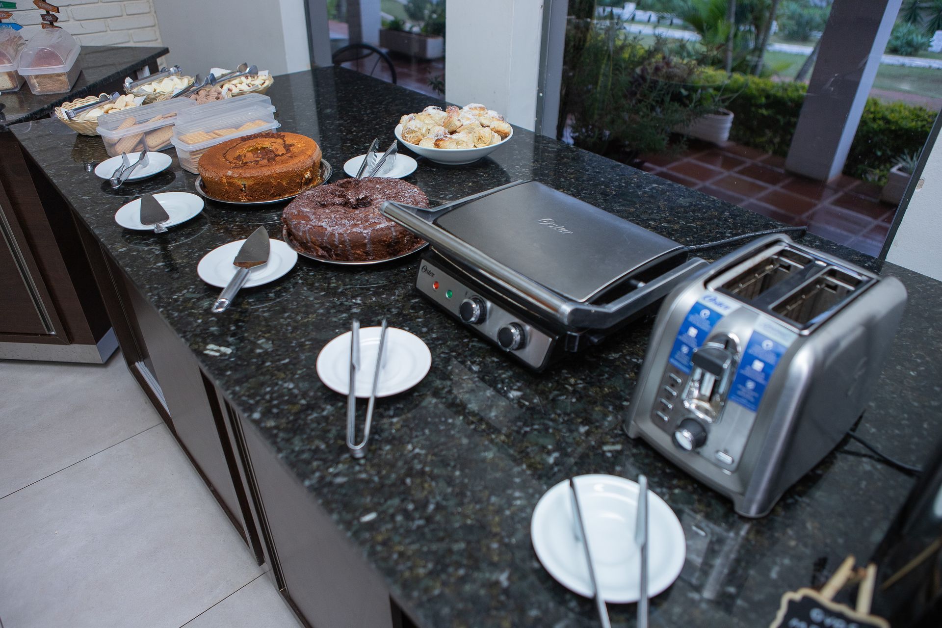 Buffet montado em bancada de granito, incluindo bolos, biscoitos, churrasqueira e torradeira.
