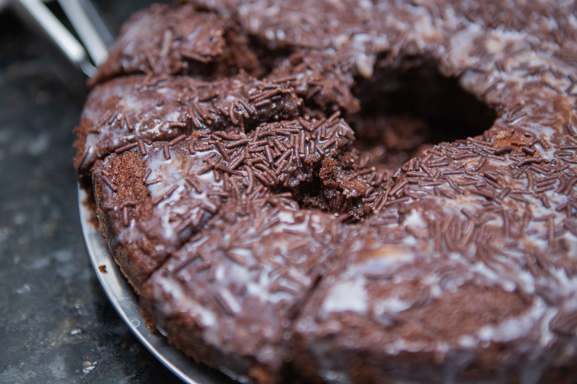Bolo de chocolate com cobertura branca e granulado, parcialmente fatiado em um prato de prata.