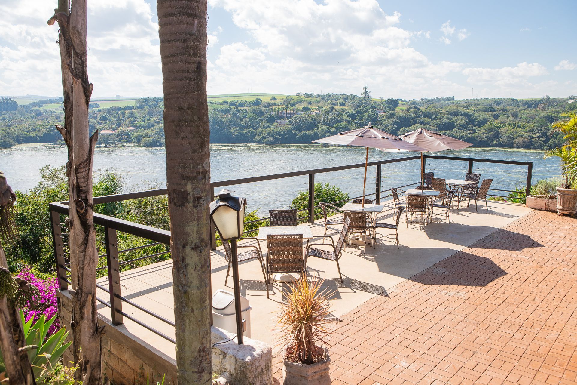 Terraço com vista para um lago, com mesas, cadeiras e um guarda-sol; árvores e céu azul visíveis.