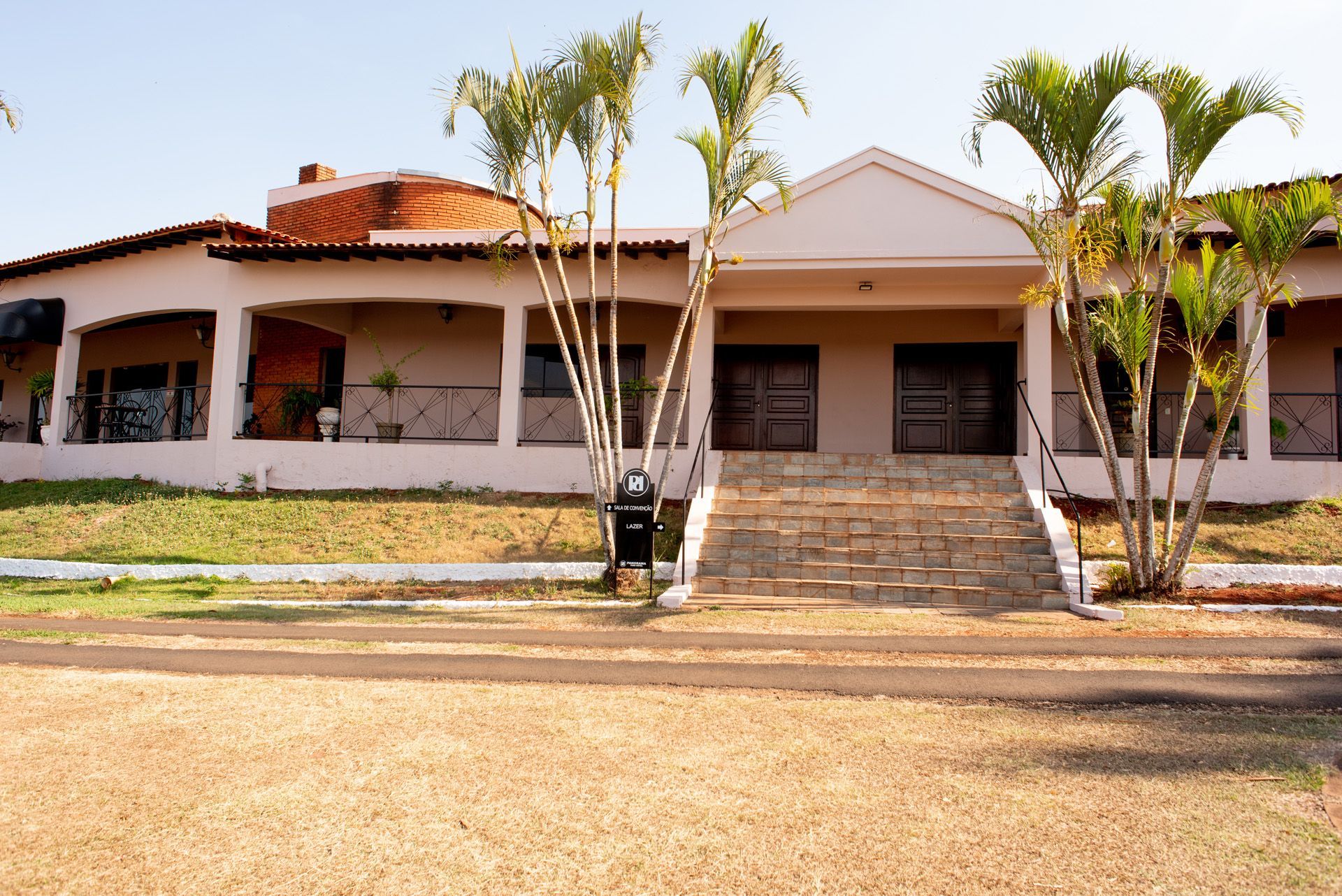 Prédio rosa de um andar com varanda coberta e degraus que levam a portas duplas, com palmeiras e um gramado.
