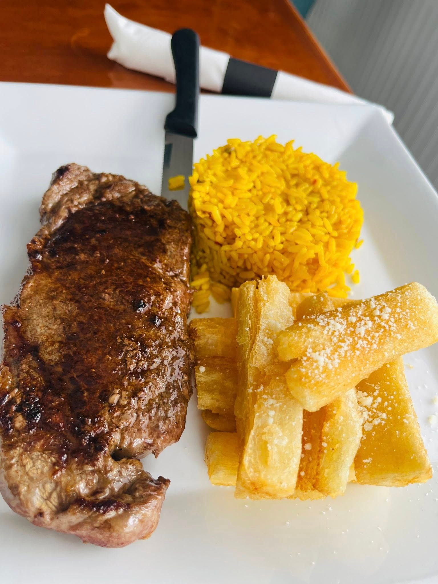 Steak, yellow rice, and fried cassava on a white plate with a knife and napkin.