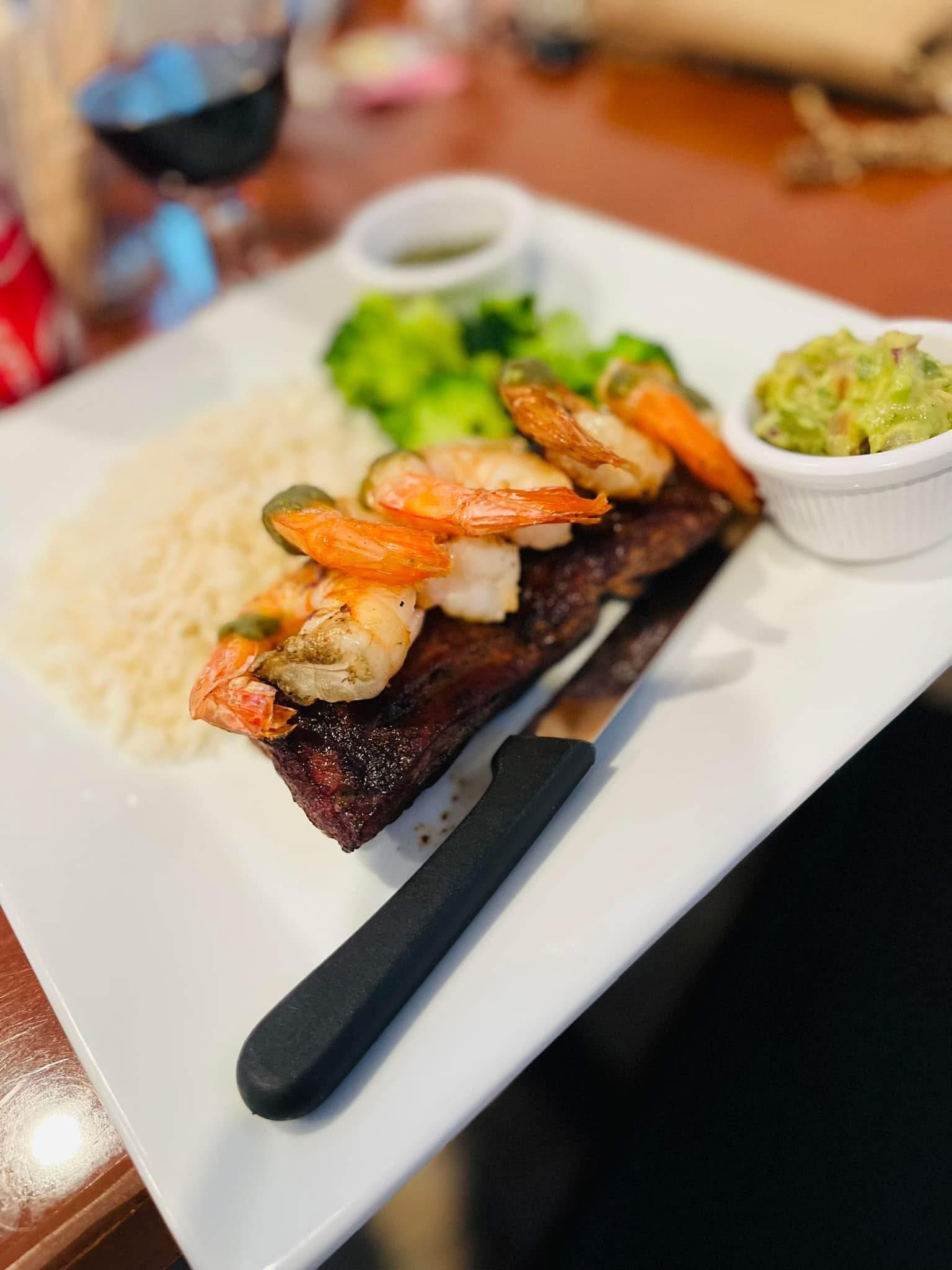 Plate of steak topped with shrimp, rice, broccoli, and guacamole; a knife rests on the plate.