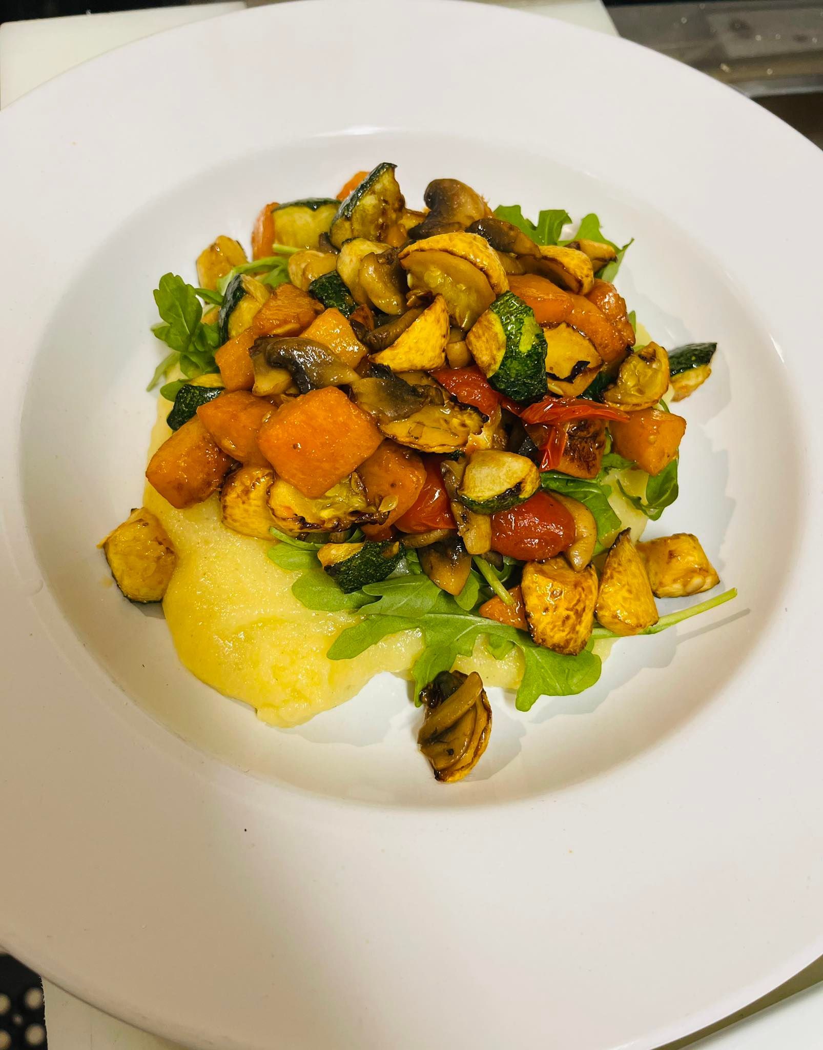 Polenta with roasted vegetables (squash, zucchini, tomatoes) and arugula on a white plate.