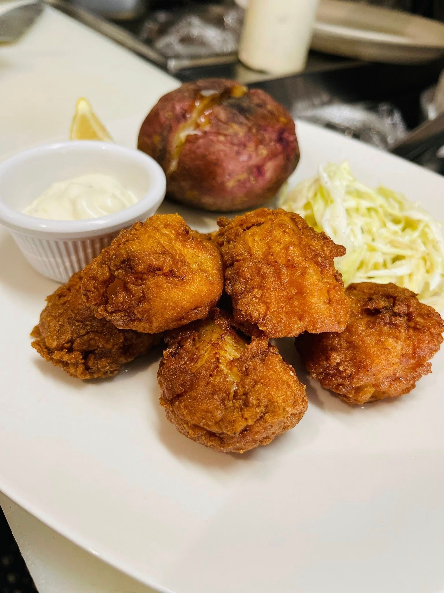 Golden fried fritters with dipping sauce, baked potato, and coleslaw on a white plate.