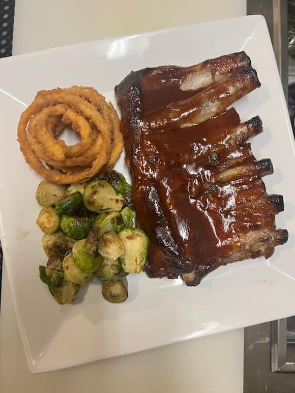 Barbecue ribs with sauce, onion rings, and roasted Brussels sprouts on a white plate.