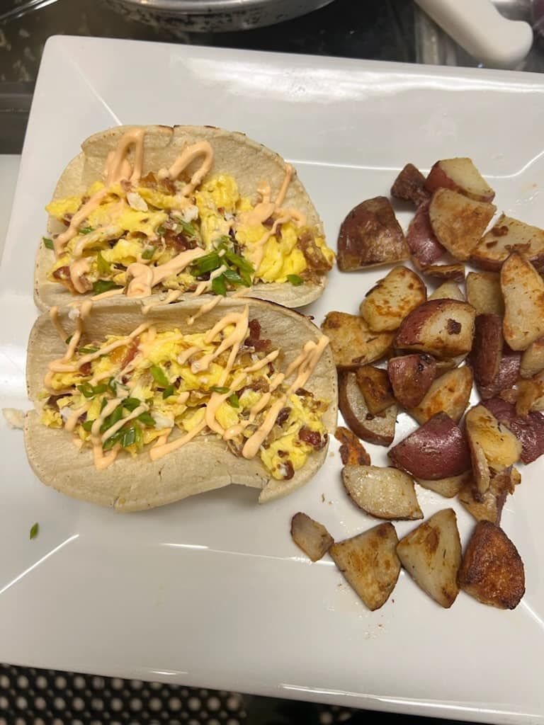 Breakfast tacos with scrambled eggs, bacon, and a side of roasted potatoes on a white plate.