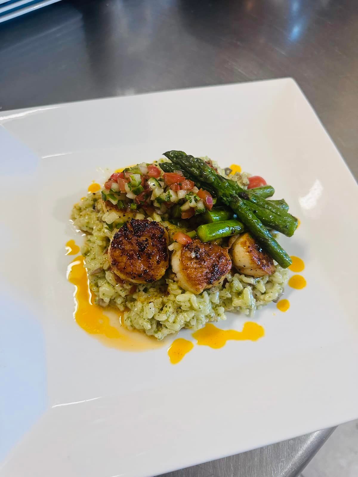 Seared scallops, pesto risotto, asparagus, and salsa on a white plate.