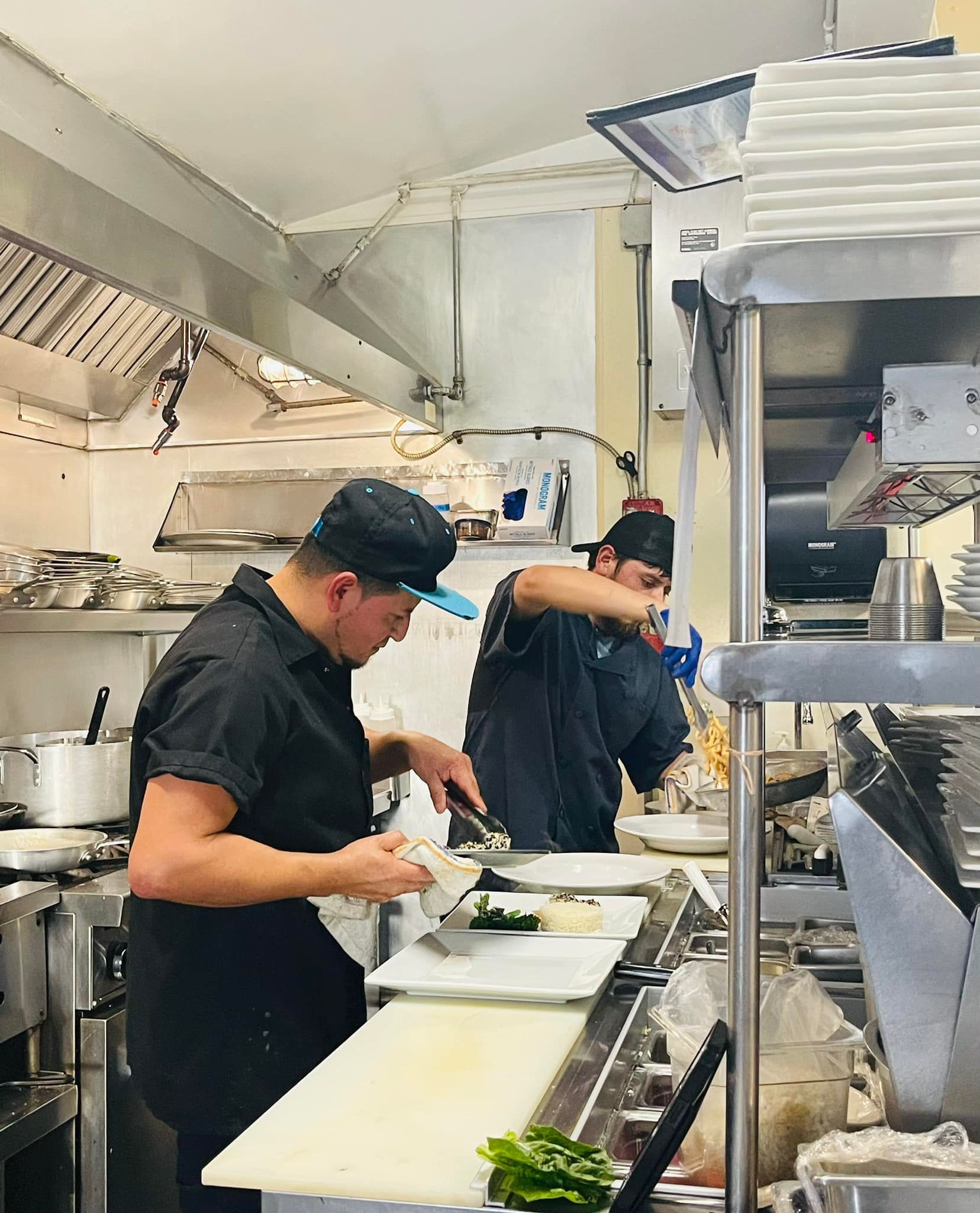 Two chefs in a busy kitchen, preparing food. One chops vegetables, the other works near a stove.