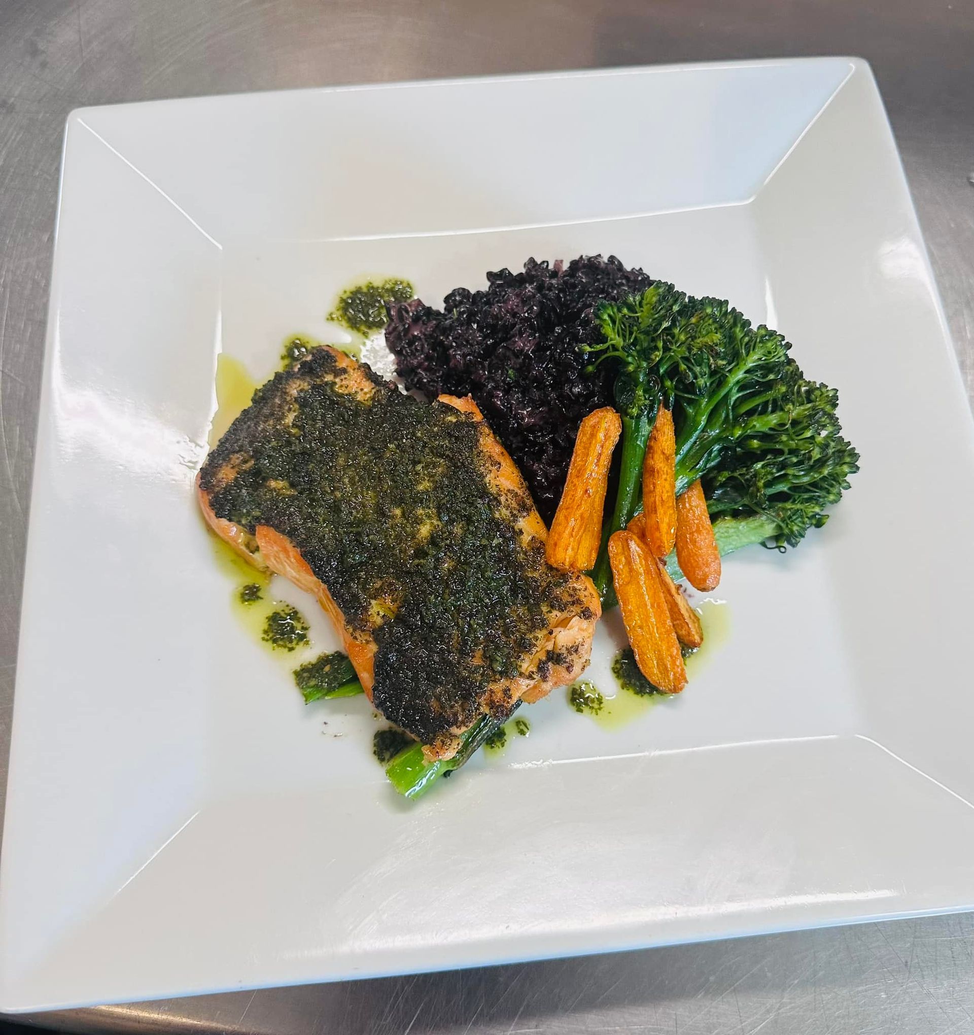 Salmon with pesto, black rice, broccoli, and carrots on a white square plate.