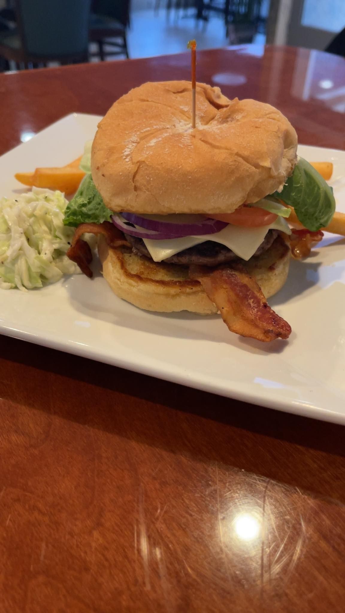 Burger with bacon, lettuce, tomato, and cheese on a white plate with coleslaw and fries.